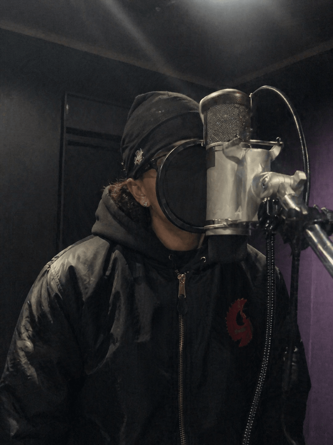 Person wearing a black hoodie and jacket, recording vocals in a studio with a microphone and pop filter.