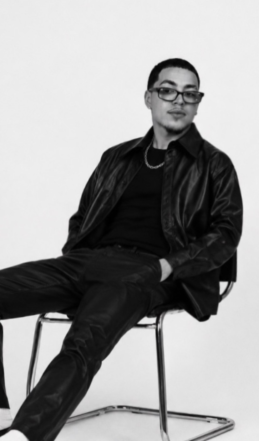 Person sitting on a chair, wearing glasses, a leather jacket, and dark clothing, against a plain background.