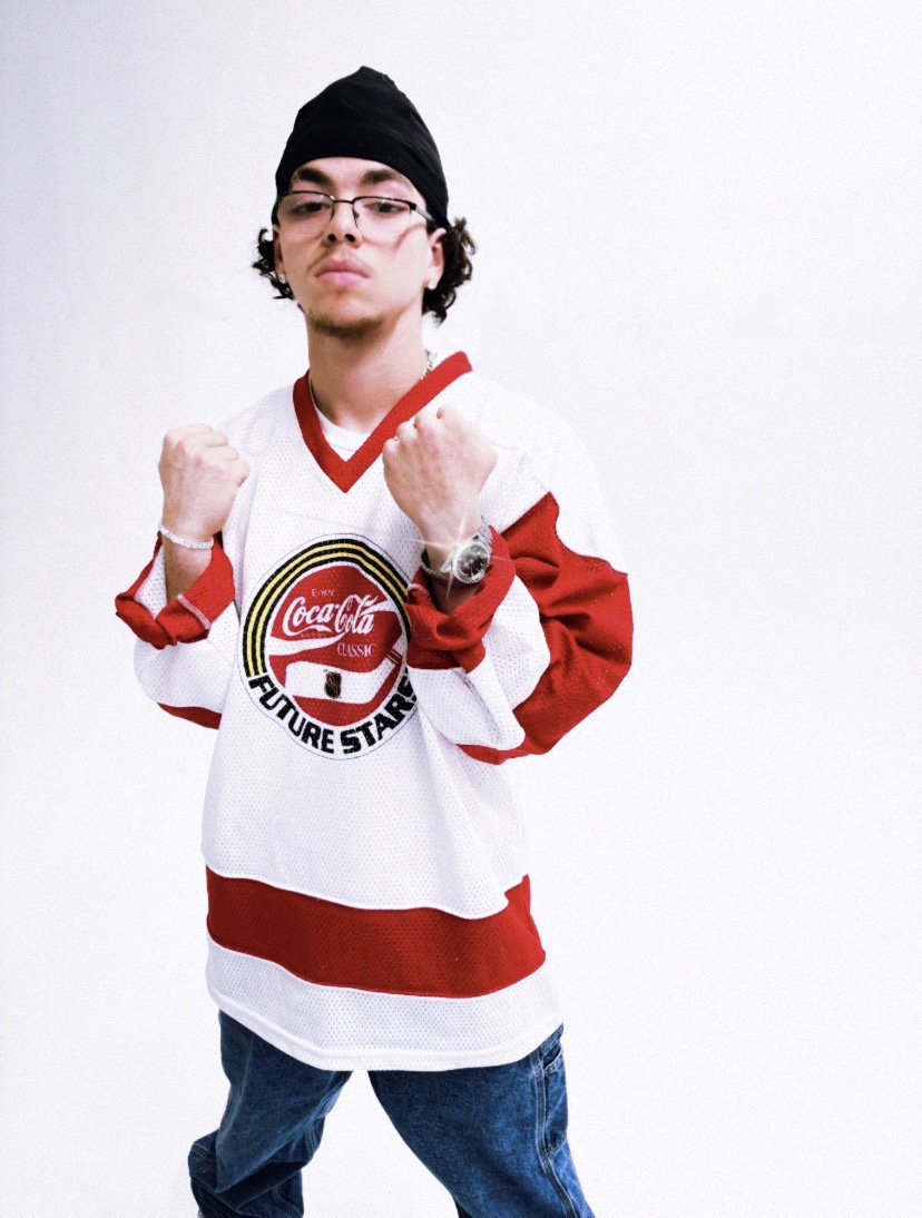 Young person wearing glasses, black beanie, and a white and red sports jersey, standing with fists raised, against a plain white background.