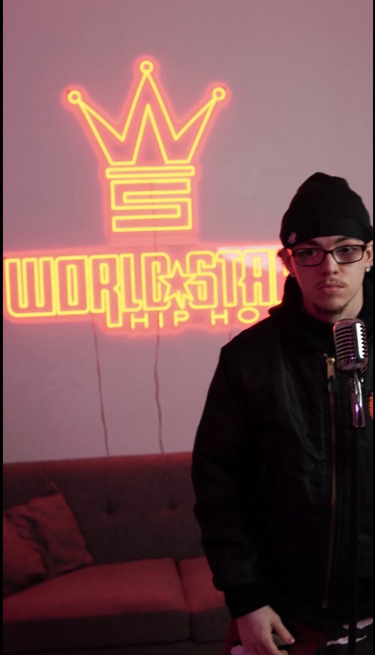 Young man wearing glasses and a black hoodie stands in front of a microphone, with a pink wall and neon sign behind him.