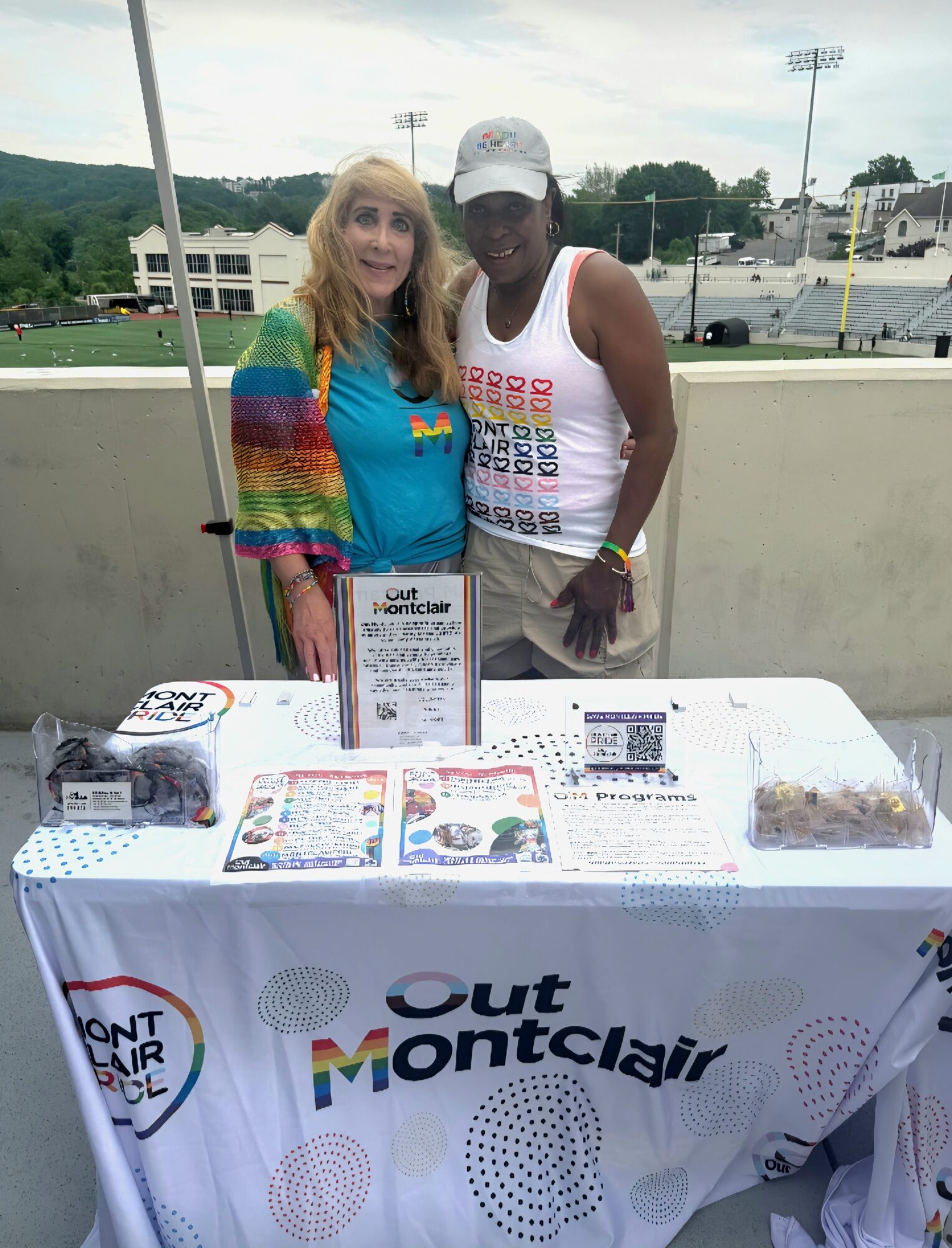 Two women stand behind a table with promotional materials outdoors, smiling at the camera.