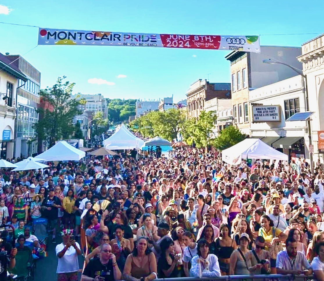 Crowd gathered on a city street with tents, buildings, and a banner overhead for Montclair Pride June 8th, 2024.