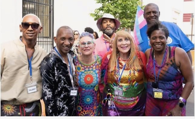 Group of seven diverse people smiling outdoors, some wearing colorful clothing and accessories, with a flag in the background.
