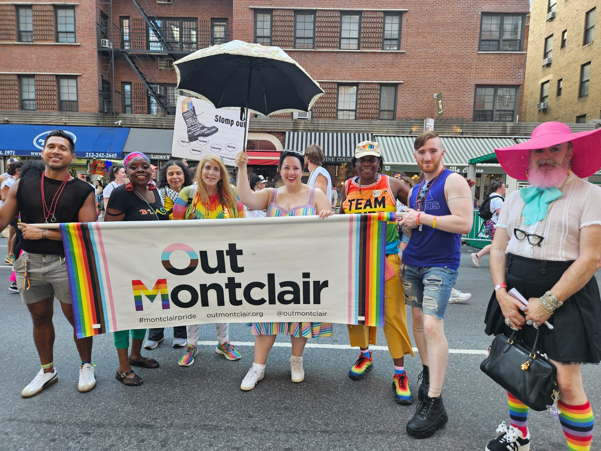 Group of people holding a banner that reads 'Out Montclair' during a parade on a city street, with rainbow decorations and buildings in the background.