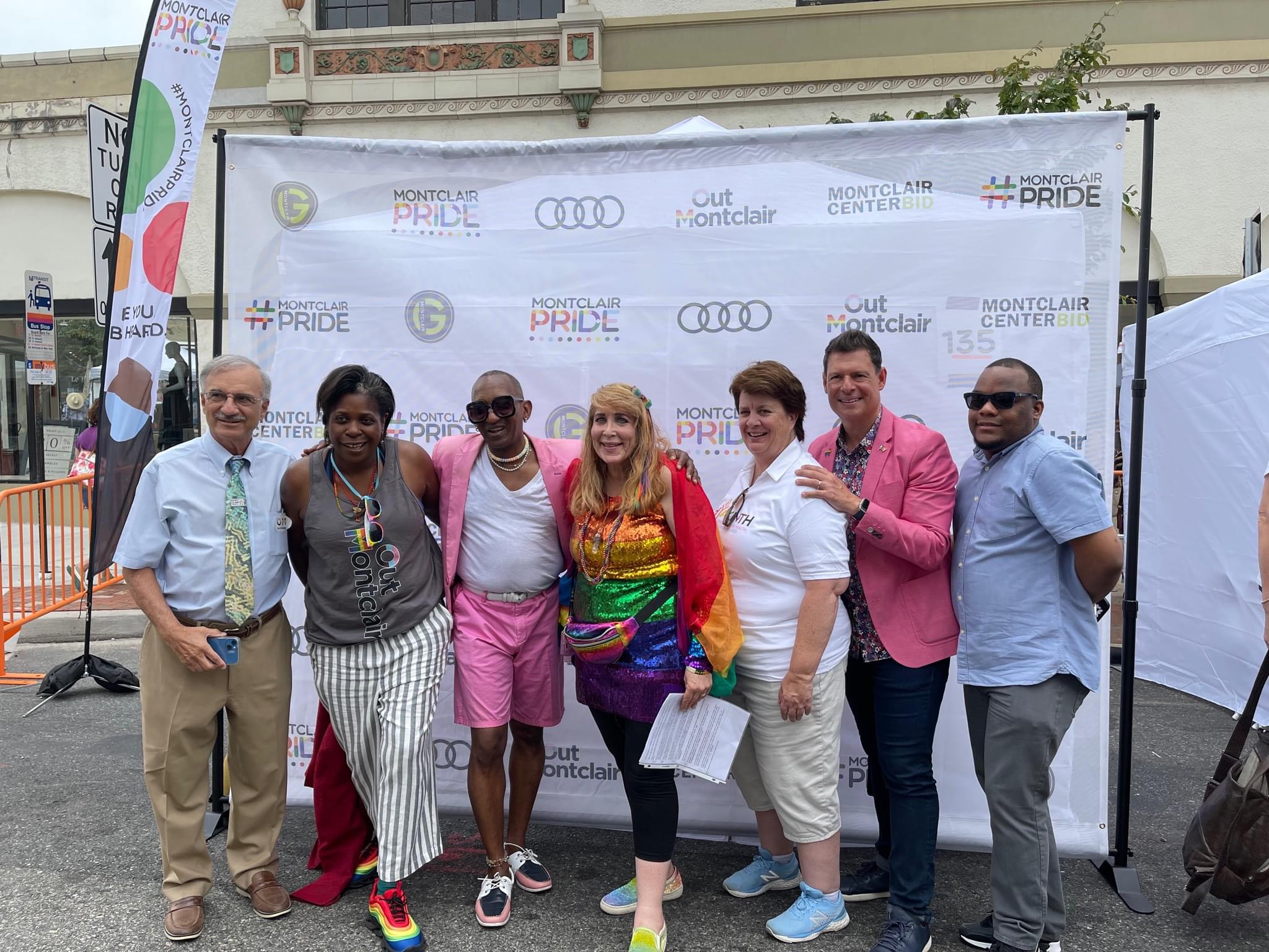 Group of seven people standing outdoors in front of a Pride event banner, smiling, with tents and flags nearby.