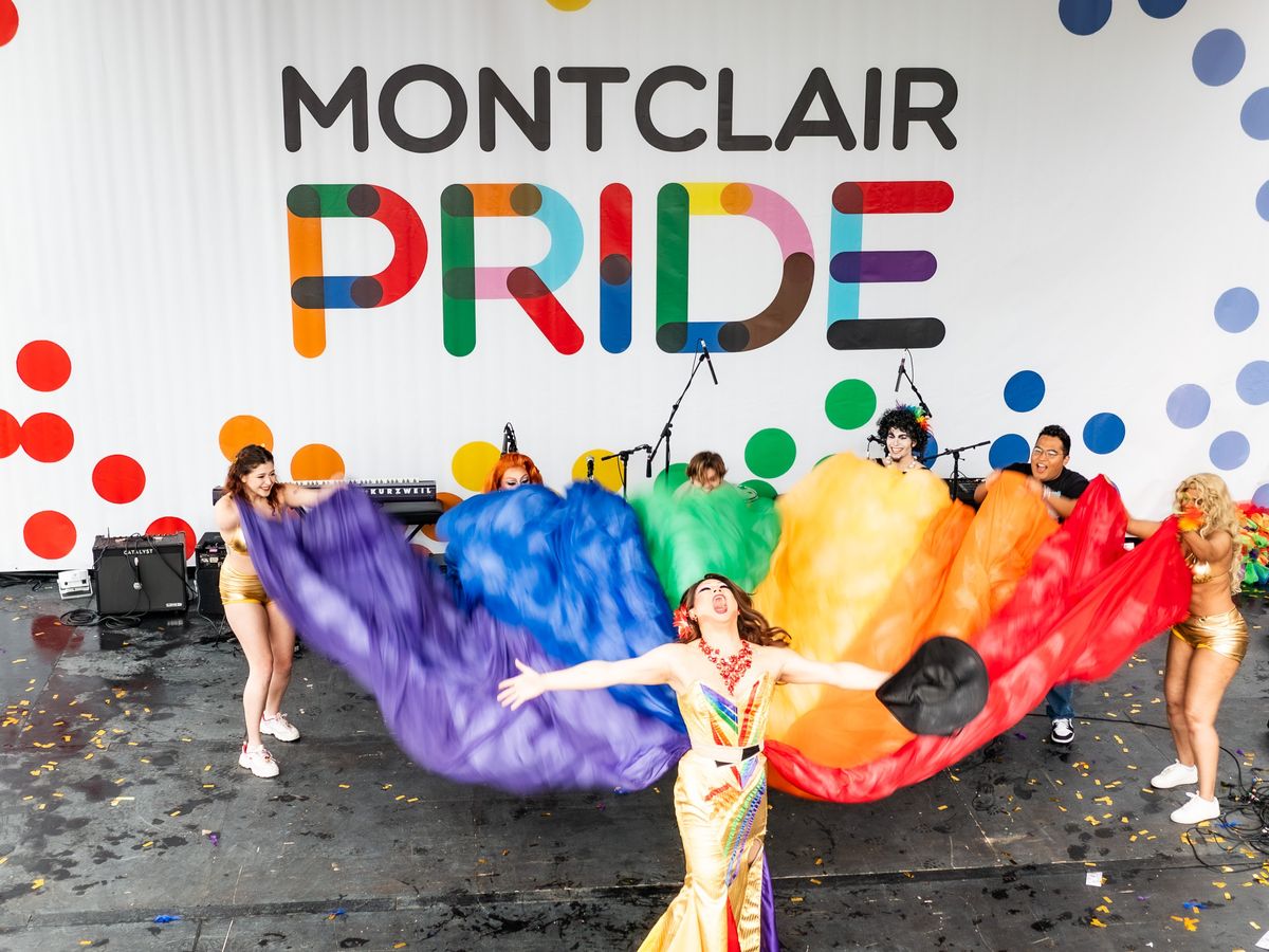 Group of people on stage holding a large rainbow-colored fabric at Montclair Pride event, with a colorful sign in the background.