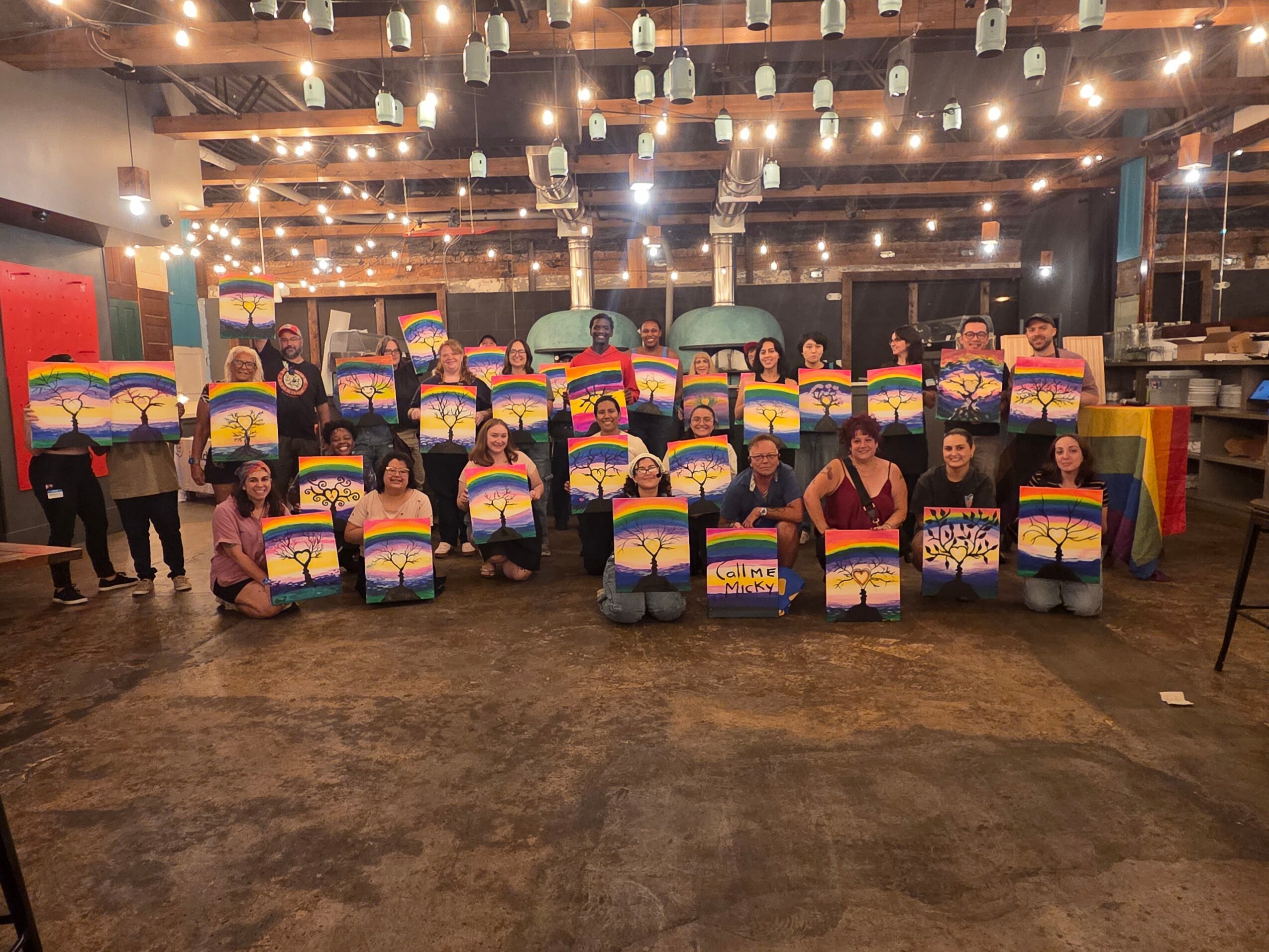 Group of people holding colorful paintings in a decorated indoor space with string lights.