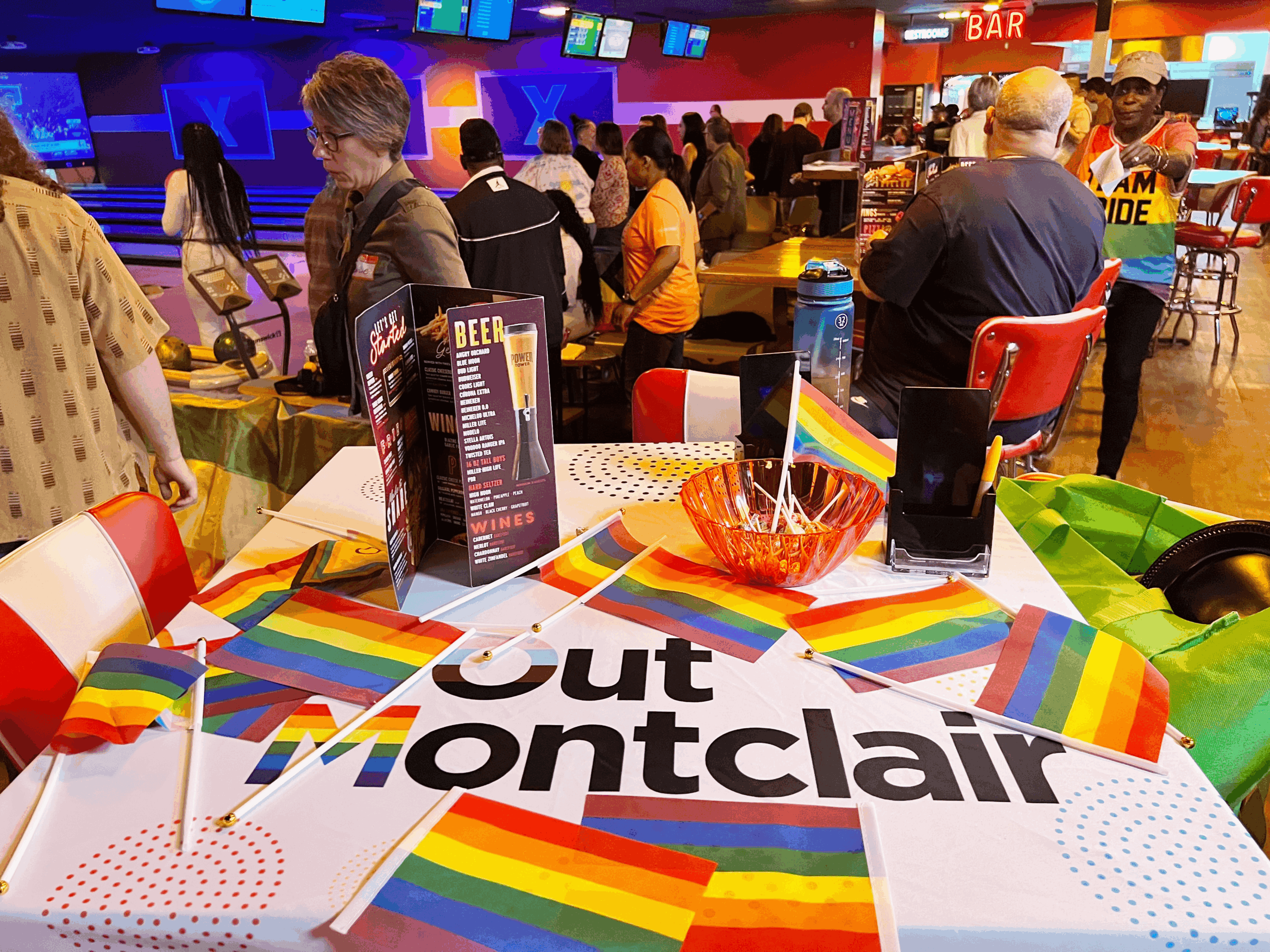 Table with rainbow decorations and a sign reading 'Montclair' at a lively bar with people in the background.
