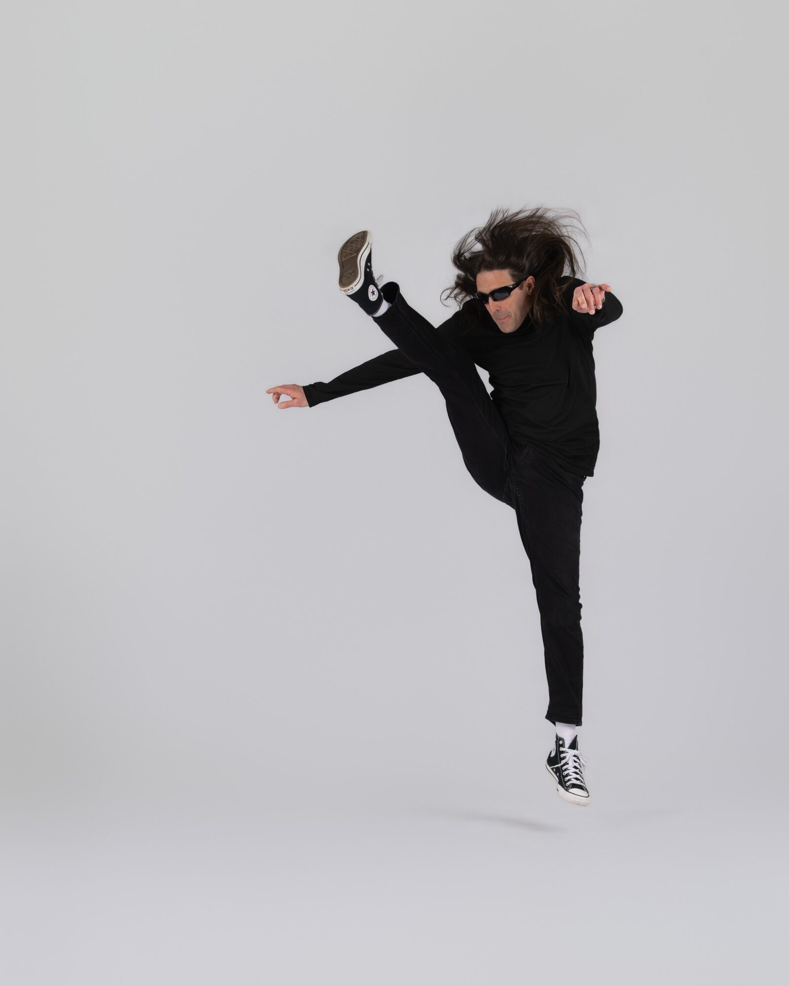 Person with long hair, sunglasses, black clothing, performing a high kick against a plain light background.