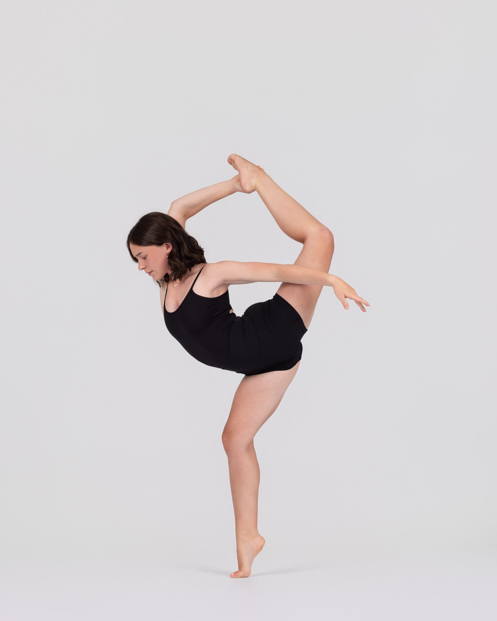 A woman in a black dress performs a dance pose with one leg raised and arms extended, against a plain background.