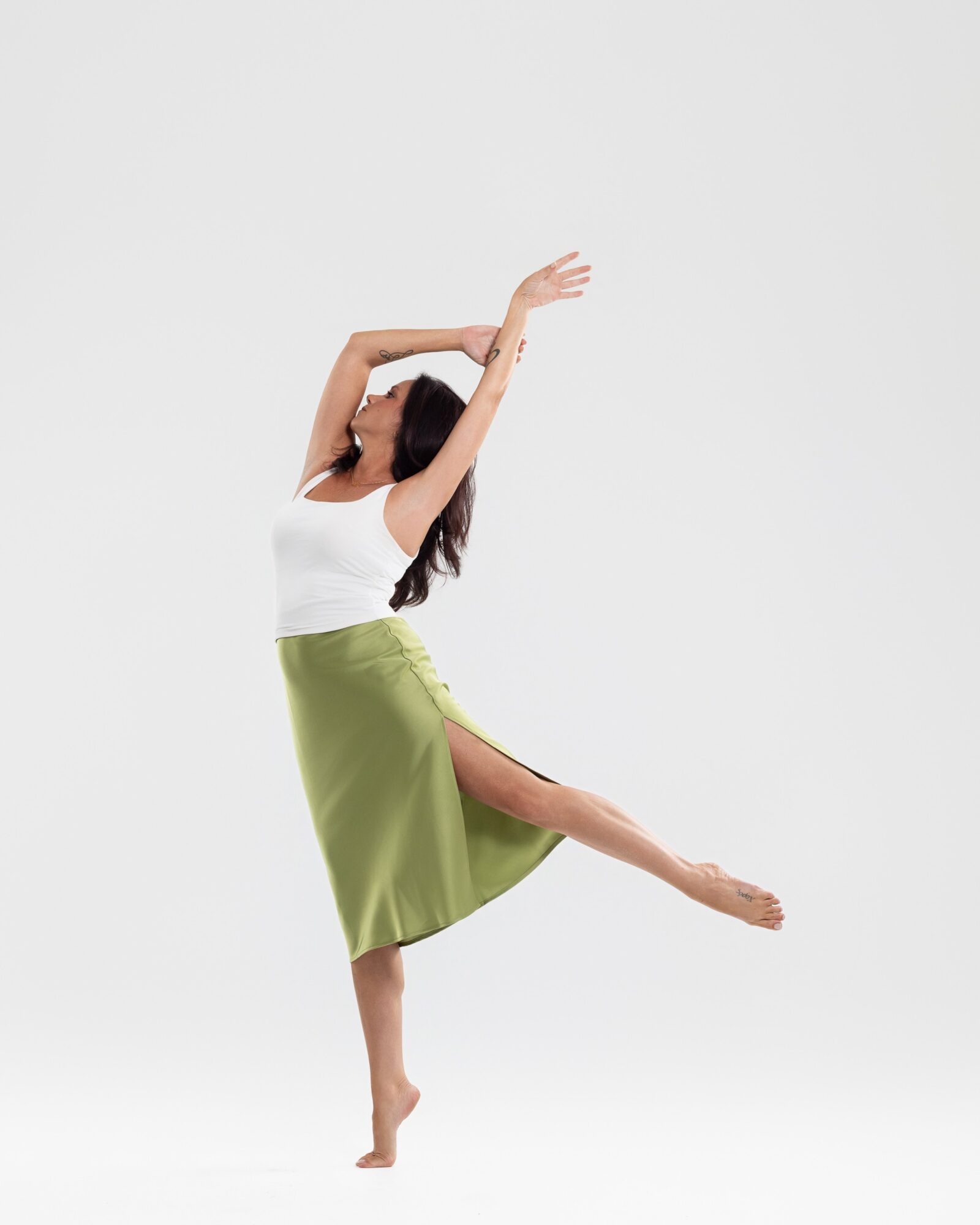 Person dancing with arms raised and one leg extended sideways, wearing a white top and green skirt, against a plain white background.