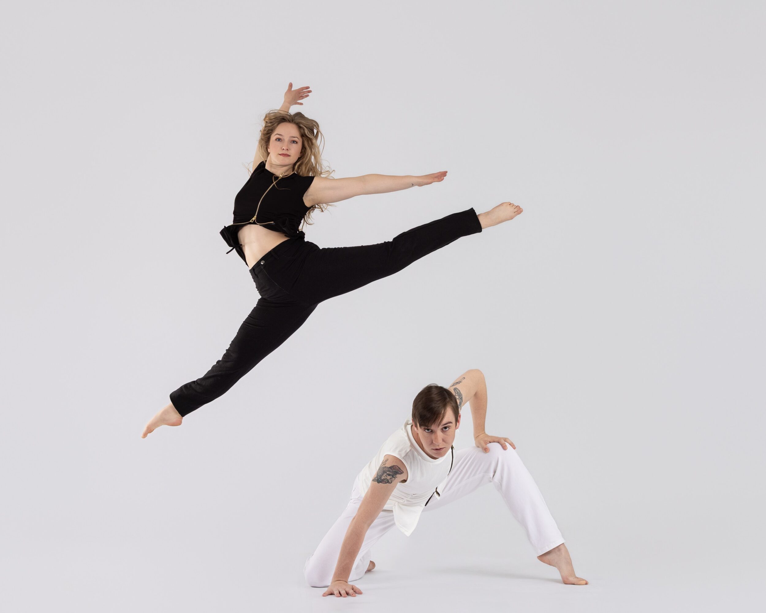 Two women perform a dance or acrobatic pose against a plain background. One is standing on hands, the other is leaping.