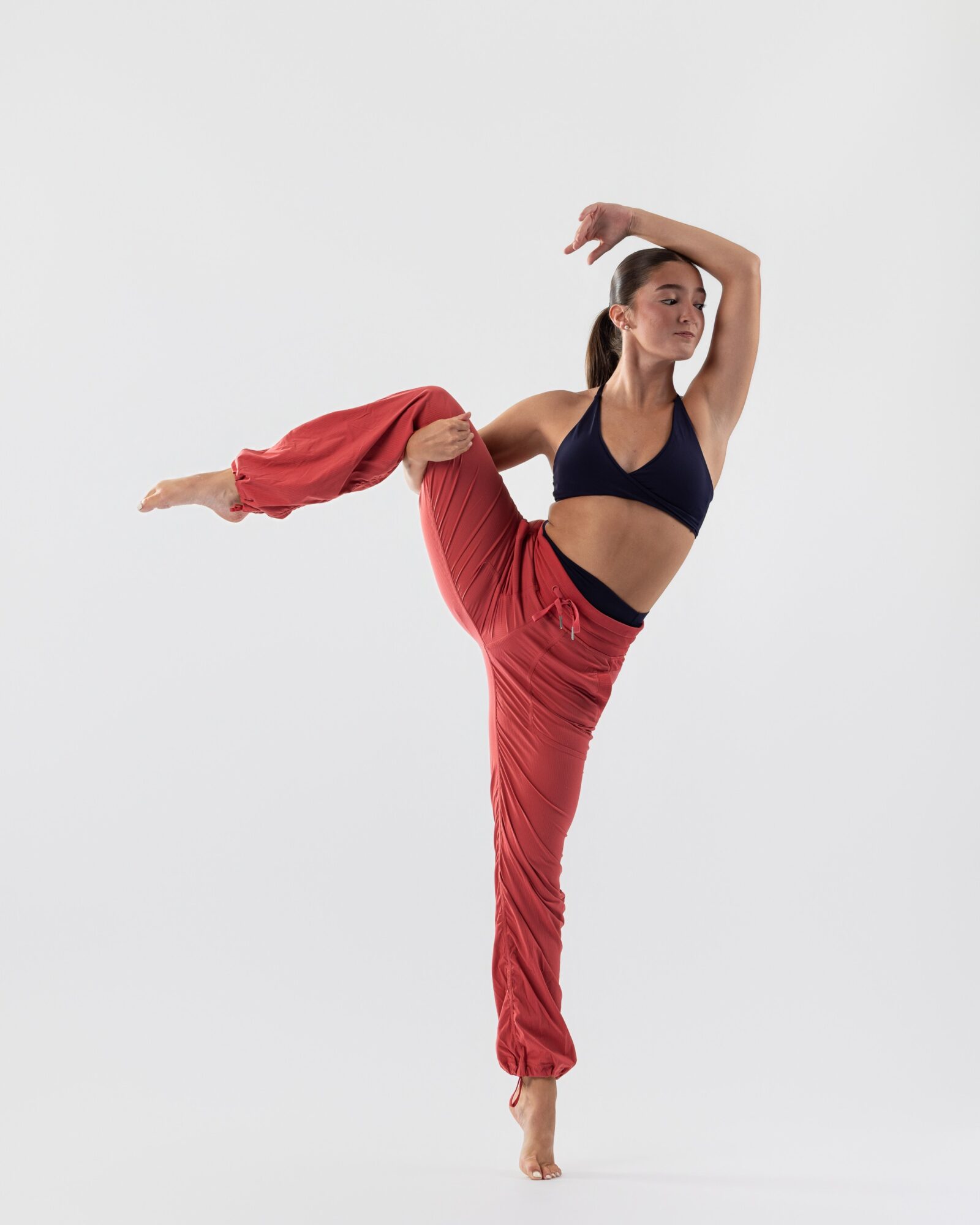 Woman in black sports bra and red pants performs a dance pose with one leg raised and arm overhead.