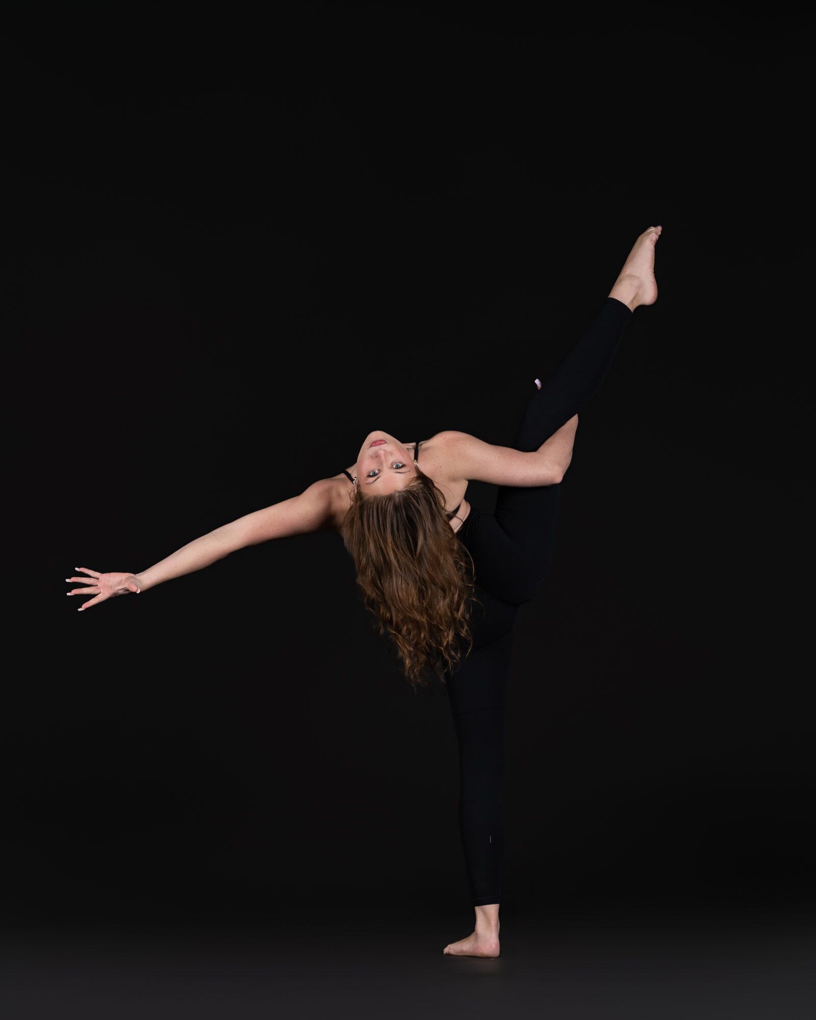 Person performing a dance pose with one leg raised and arms extended, against a black background.