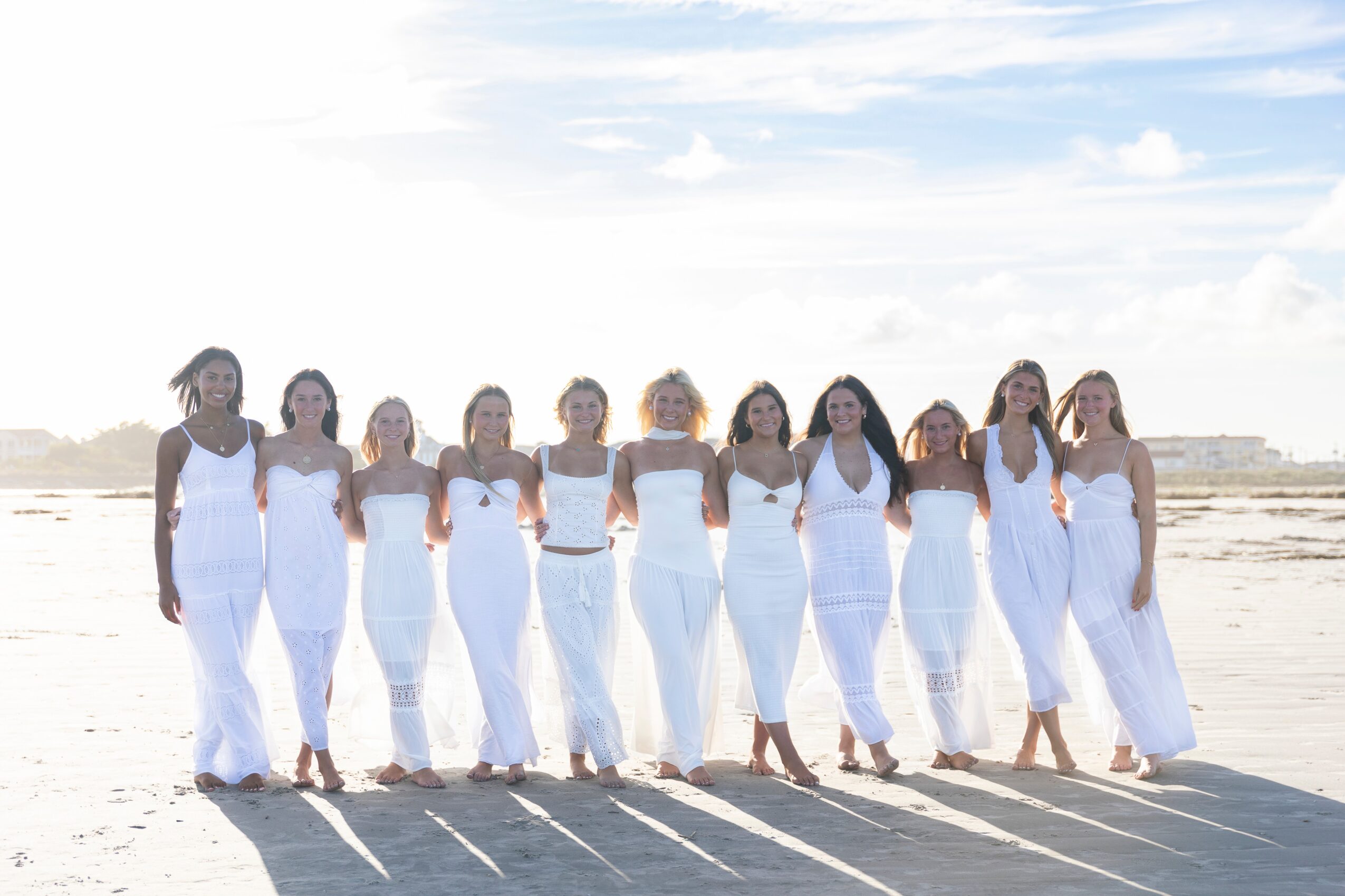 Group of ten women in white dresses standing on a beach, facing forward, with shadows cast on sand, under a partly cloudy sky.