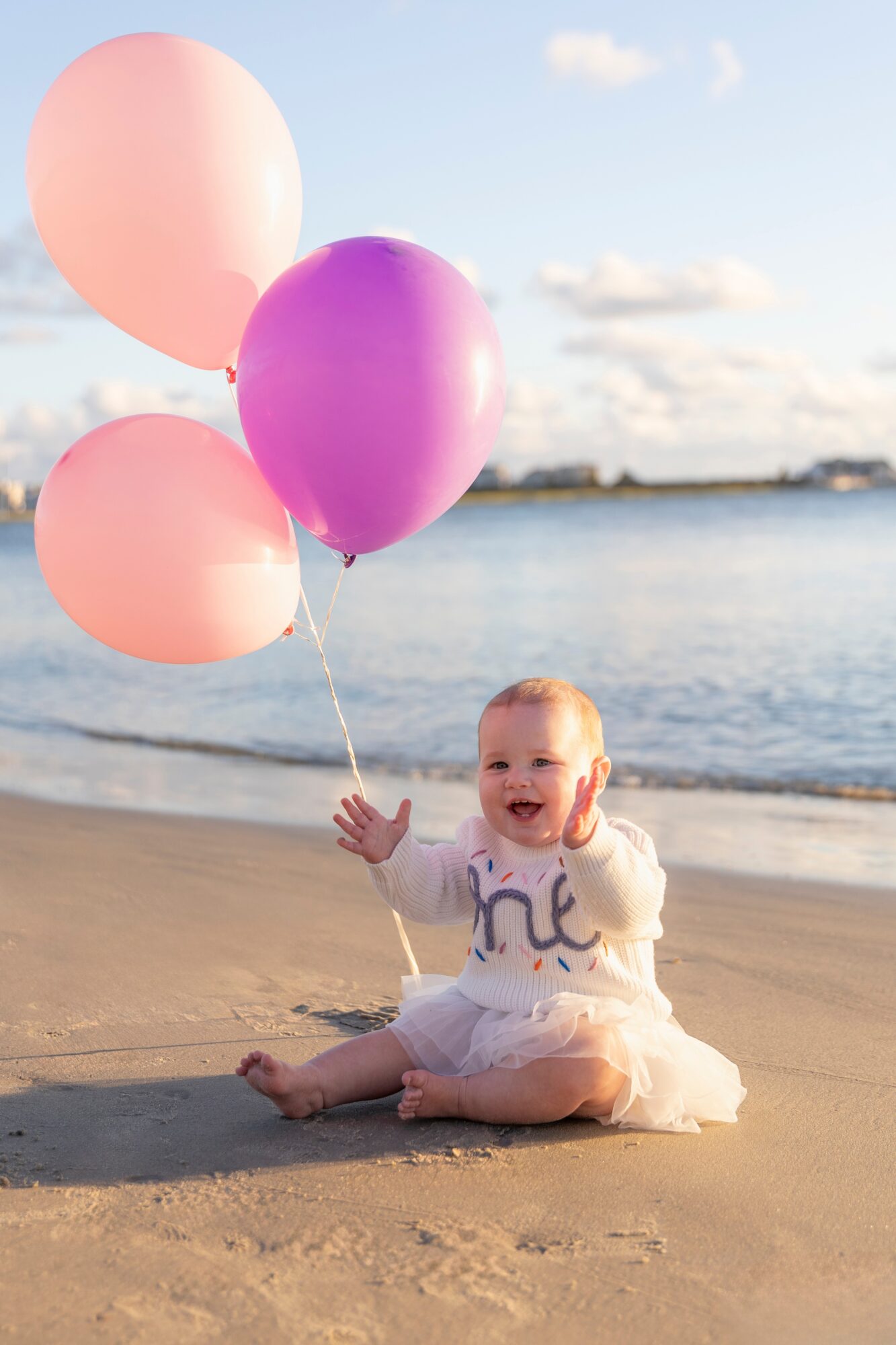 Baby sitting on sand holding three pink and purple balloons near water, smiling, with sky and clouds in background.