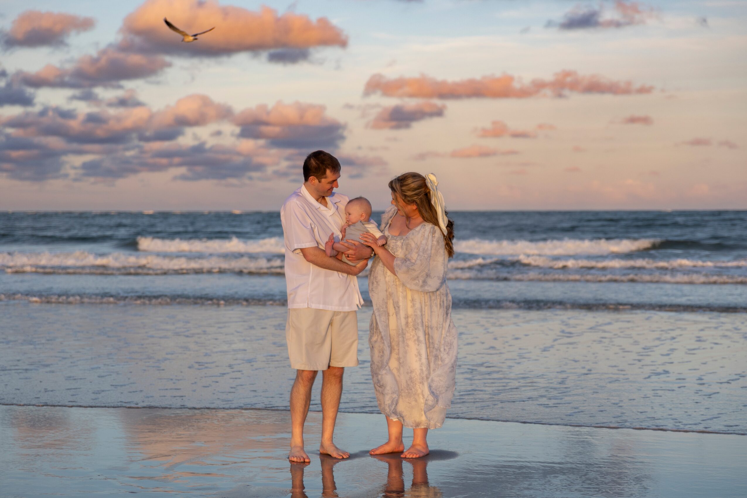 Family of three on the beach at sunset, with ocean waves and a bird flying in the sky, parents holding their baby.
