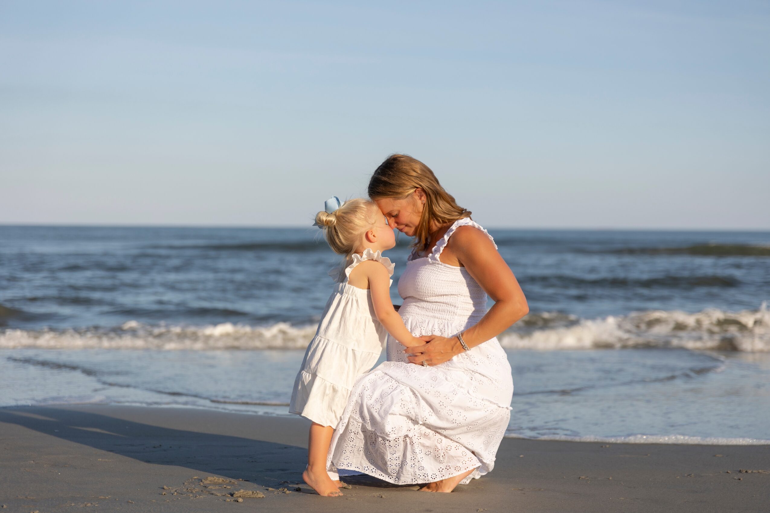 Woman kneeling on the beach with a young girl, both touching foreheads, ocean waves in background, sunny day.