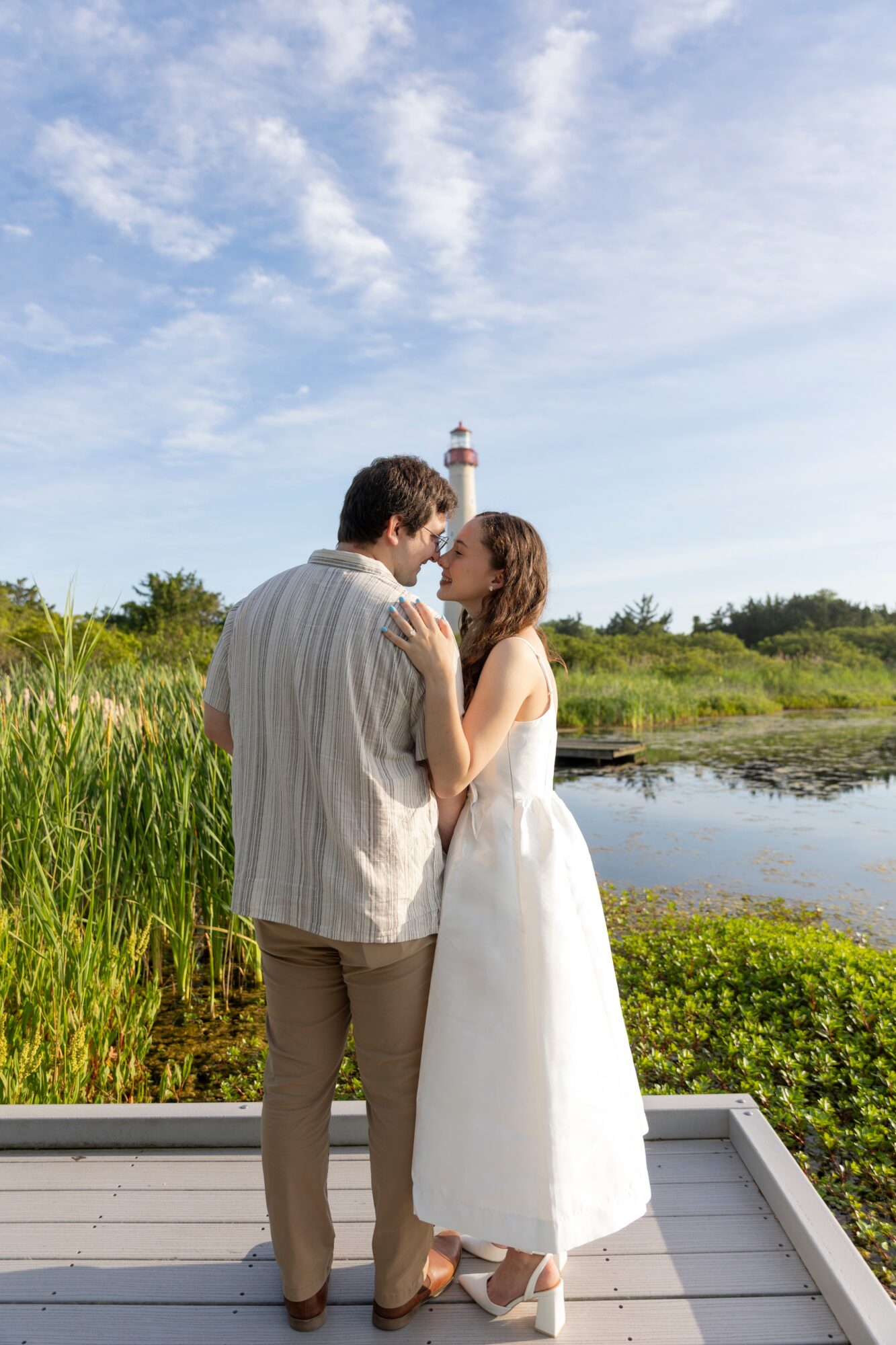 A couple stands on a deck near a pond, embracing with a lighthouse and blue sky in the background.