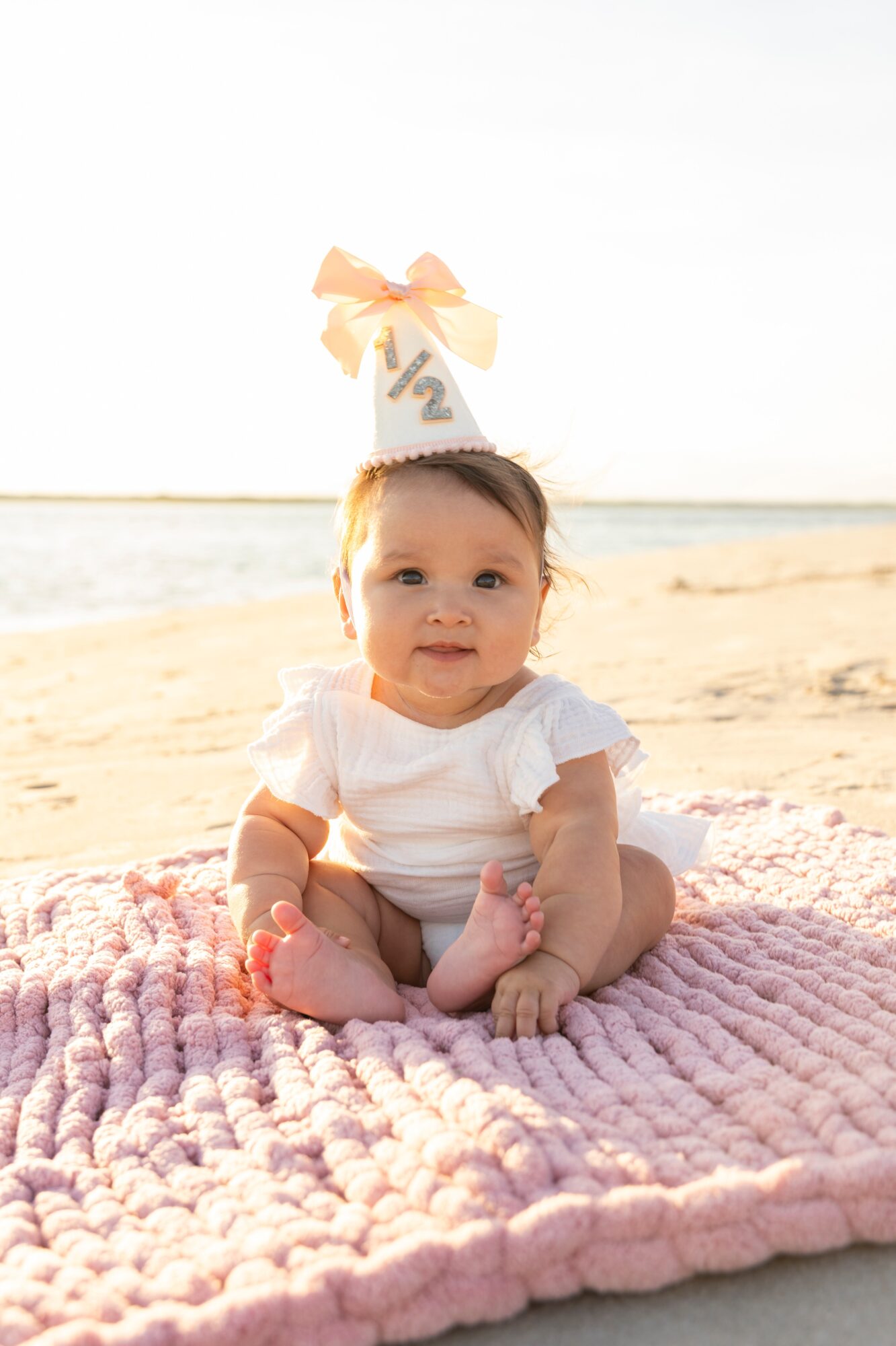 Baby sitting on a pink blanket at the beach with a birthday hat and a half-year sign, sunny sky, ocean in background.