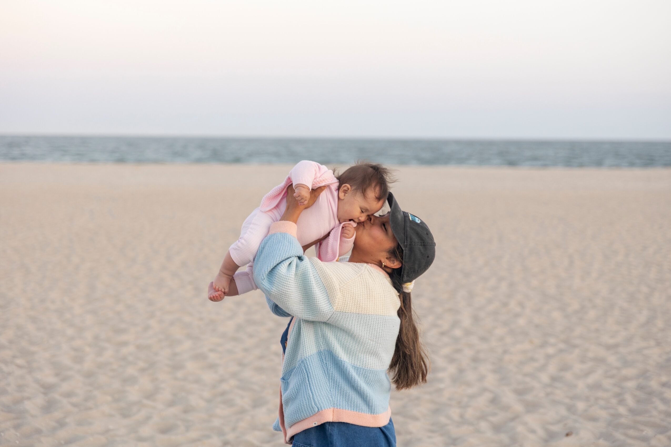 Person holding a child on a beach with sand and ocean in the background, sky above, and both facing each other.