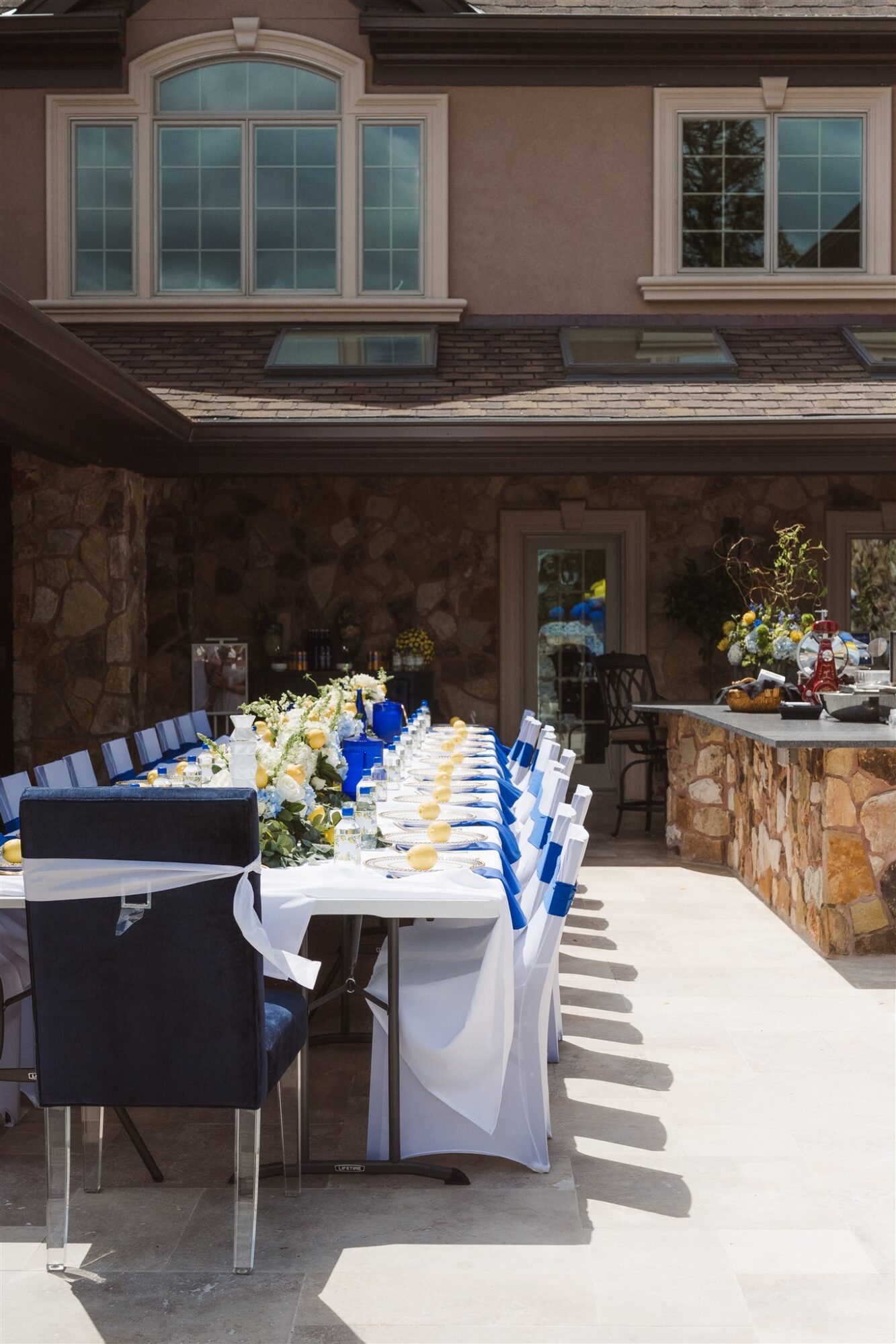 Outdoor dining table set with white tablecloth, blue and white chairs, and floral centerpiece, in front of a house with large windows.
