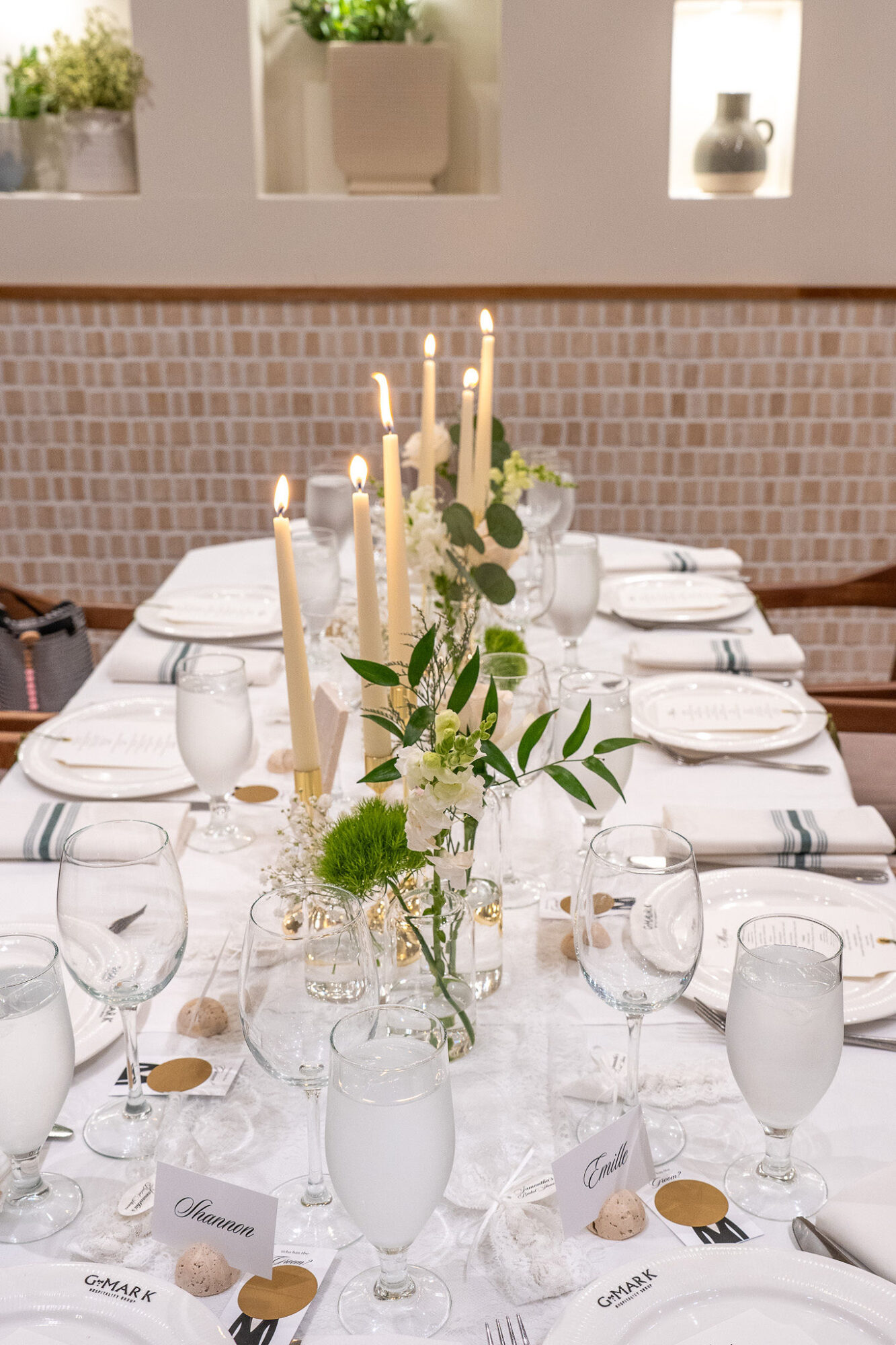 Elegant table setting with white tablecloth, candles, glassware, and floral centerpiece in a bright room.