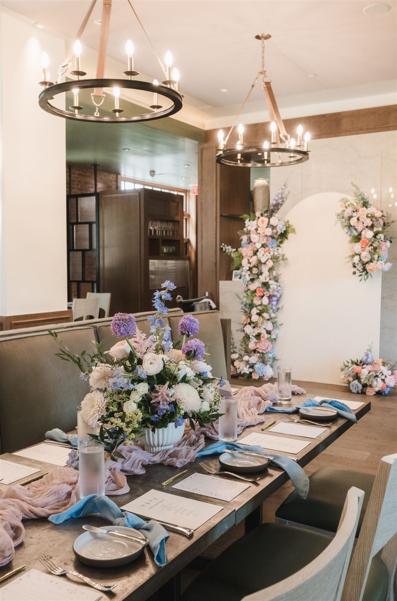 Long dining table with floral centerpieces, set with plates, napkins, and glasses, in a decorated room with floral arches and chandeliers.