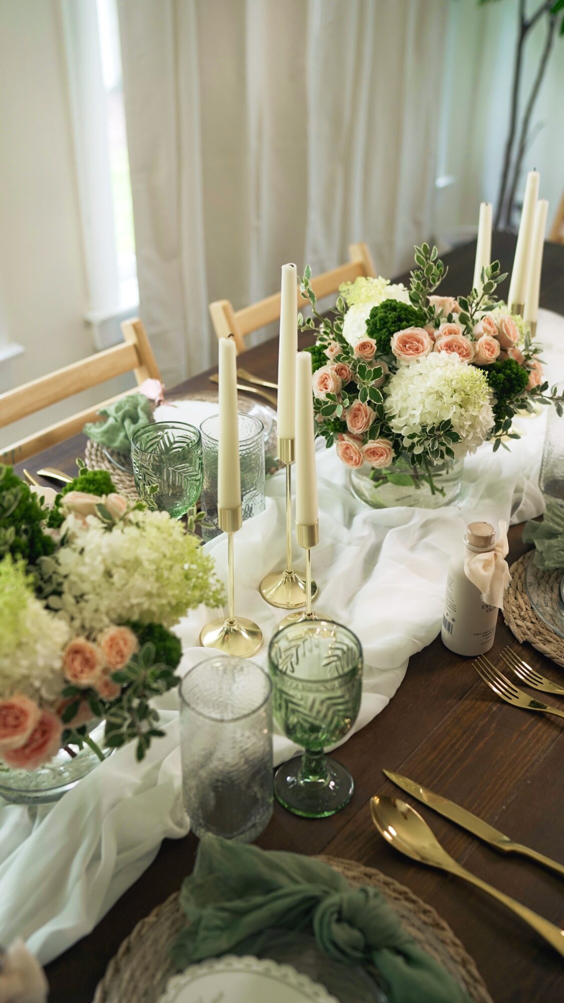 Elegant table setting with floral arrangements, candles, and gold utensils on a white tablecloth, in a bright room.