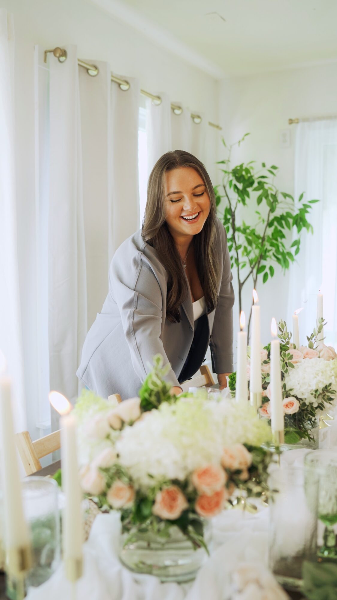 Woman smiling at a table with floral arrangements in a bright room with white curtains and a large plant.