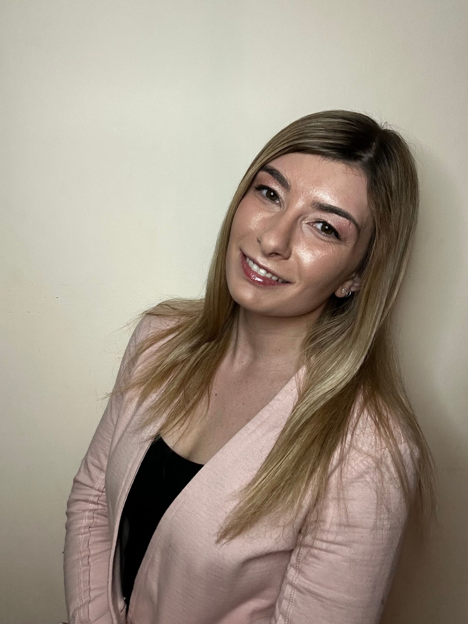 Young woman with long hair smiling, wearing a light pink blazer over a black top, standing against a plain wall.