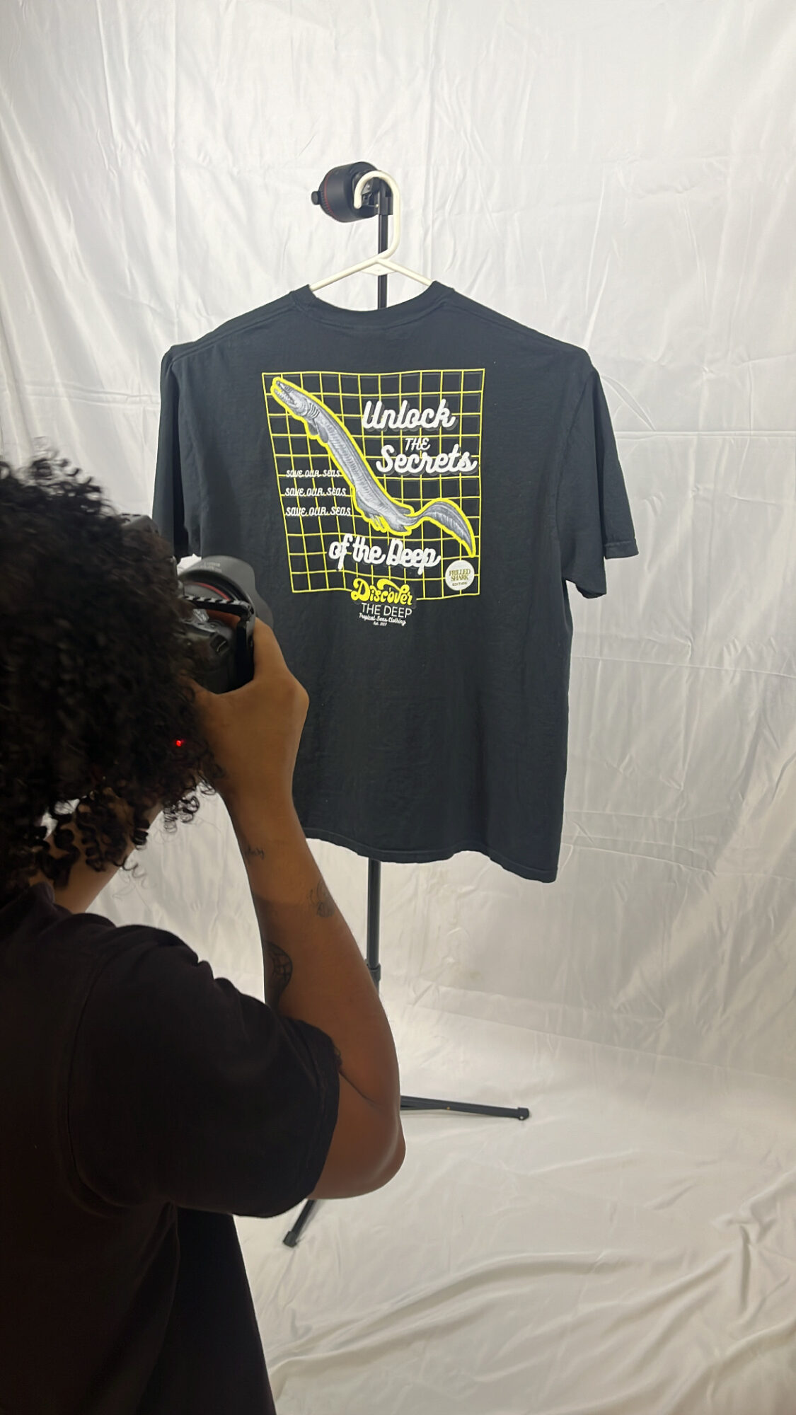 Person taking a photo of a black T-shirt hanging on a stand, with a graphic design and text, against a white background.