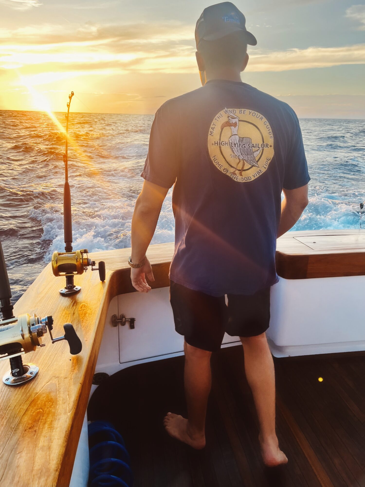 Person standing on a boat facing the ocean during sunset, with fishing equipment nearby.