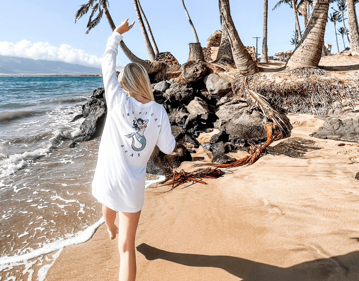 Person with blonde hair in a white dress walking on a sandy beach near palm trees and rocks, ocean in background.