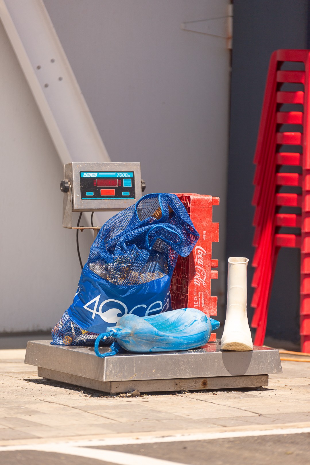 Blue plastic bag with shoes and a red container on a weighing scale, against a gray wall with stacked red chairs nearby.