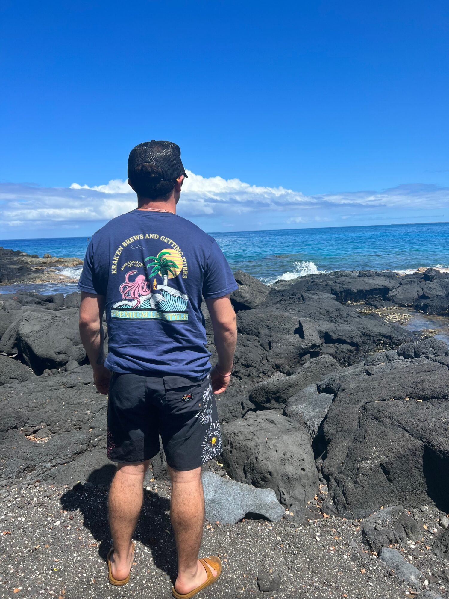 Person standing on rocky shoreline facing the ocean under a blue sky with clouds.