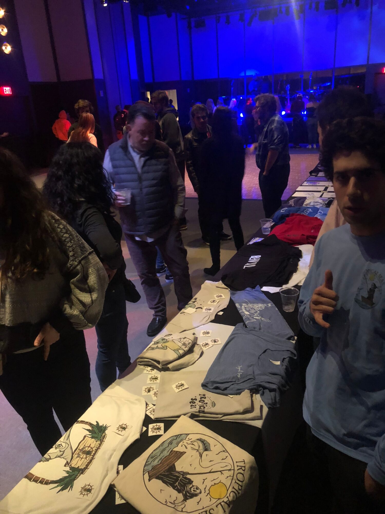 Group of people gathered around tables with artwork and posters, in a dimly lit room with blue lighting.