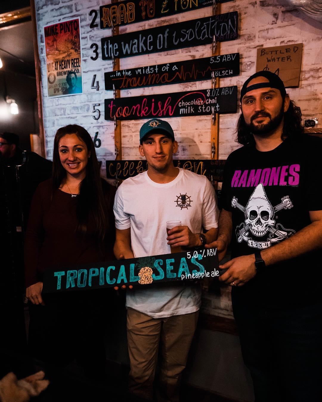 Three people standing indoors, holding a sign that reads 'TROPICAL SEA.' Behind them is a menu board with various items listed.