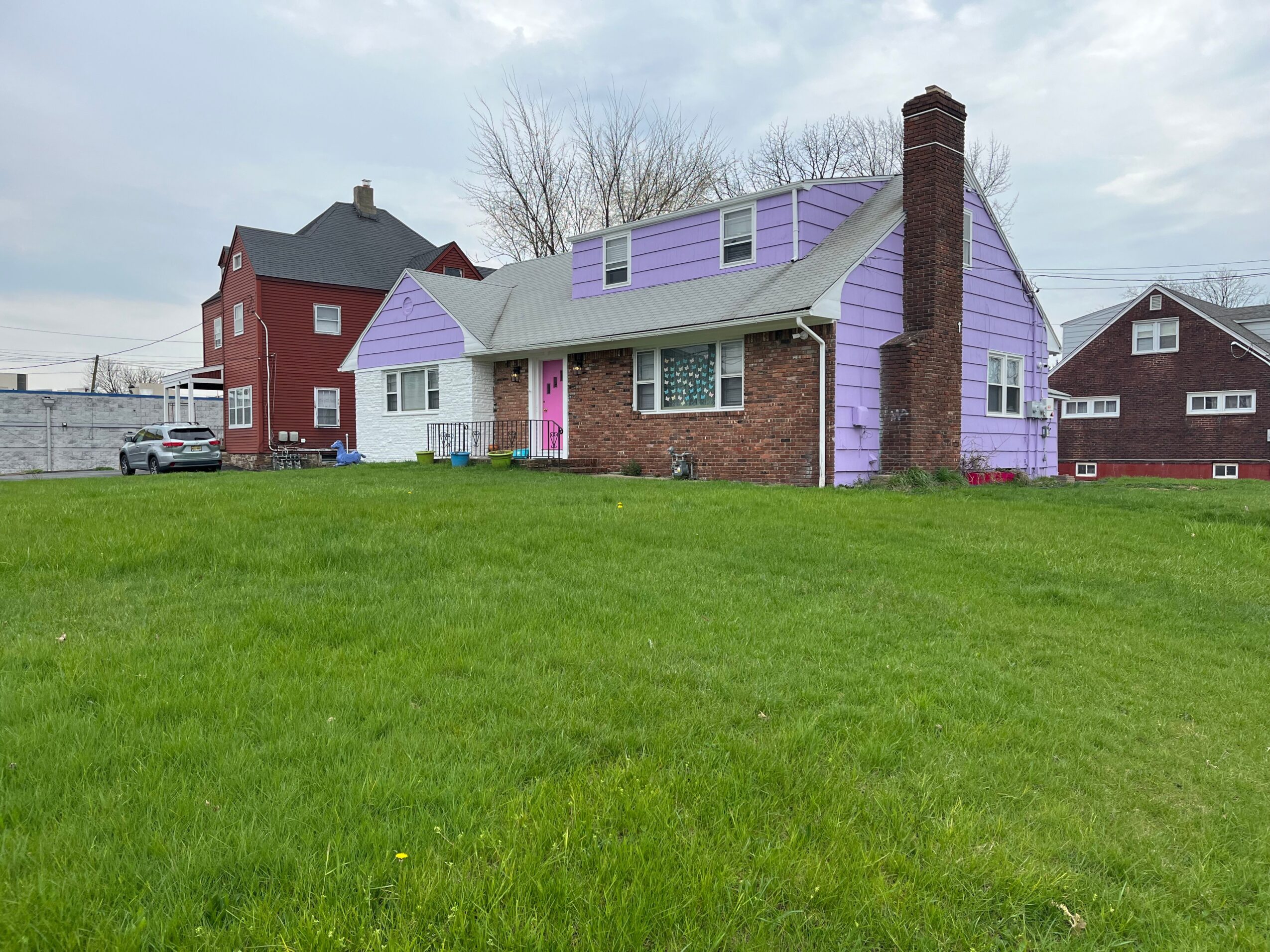 House with purple siding, brick foundation, and chimney, surrounded by a grassy lawn and neighboring houses, under a cloudy sky.