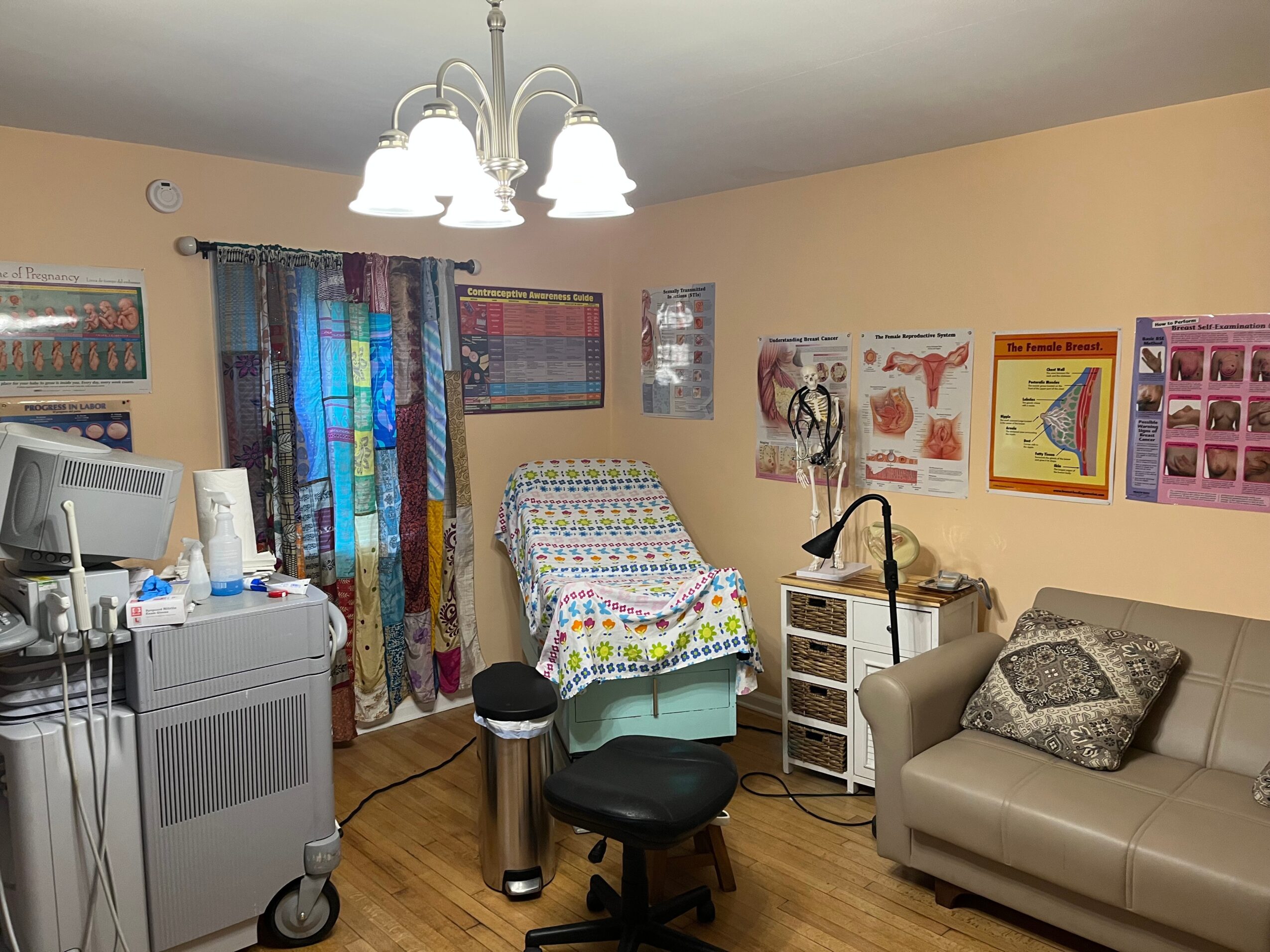 Medical examination room with a bed, medical posters, a sofa, and medical equipment, illuminated by ceiling lights.