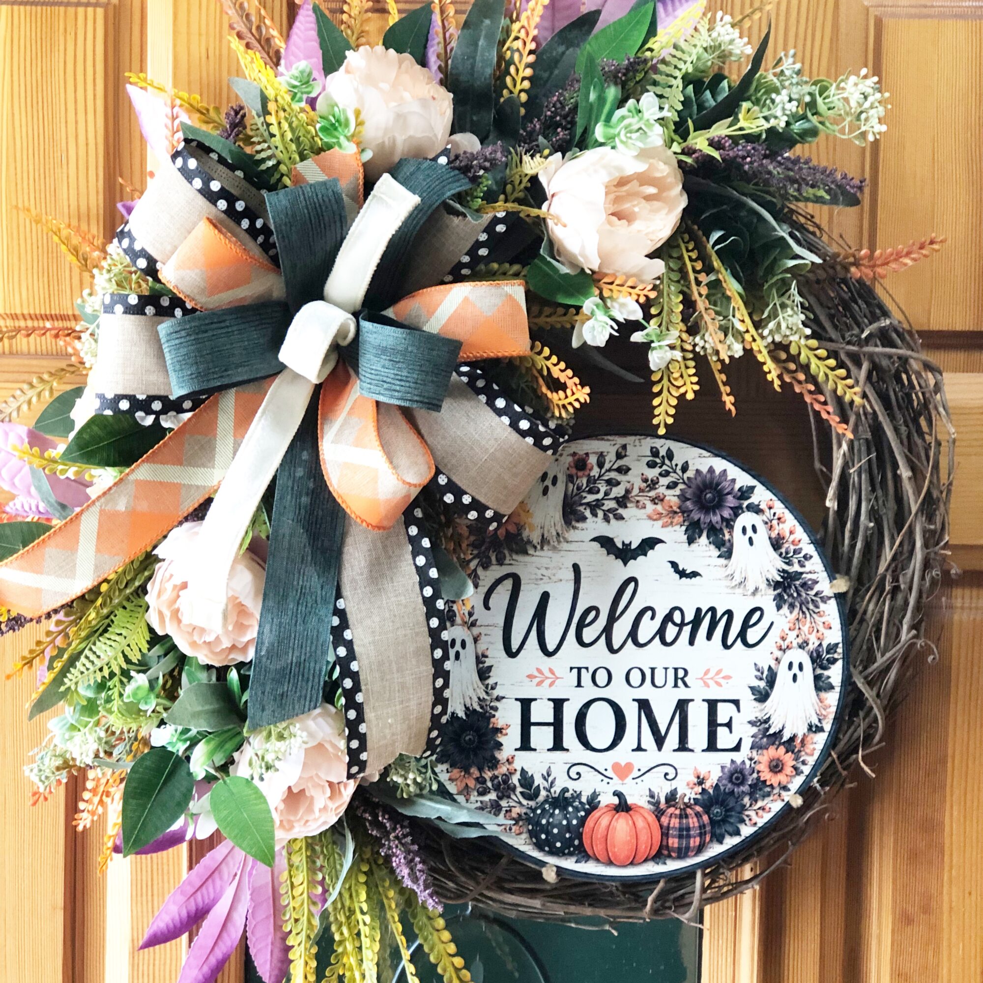 Decorative wreath with flowers, ribbons, and a sign reading 'Welcome to our home' with pumpkins, on a wooden background.