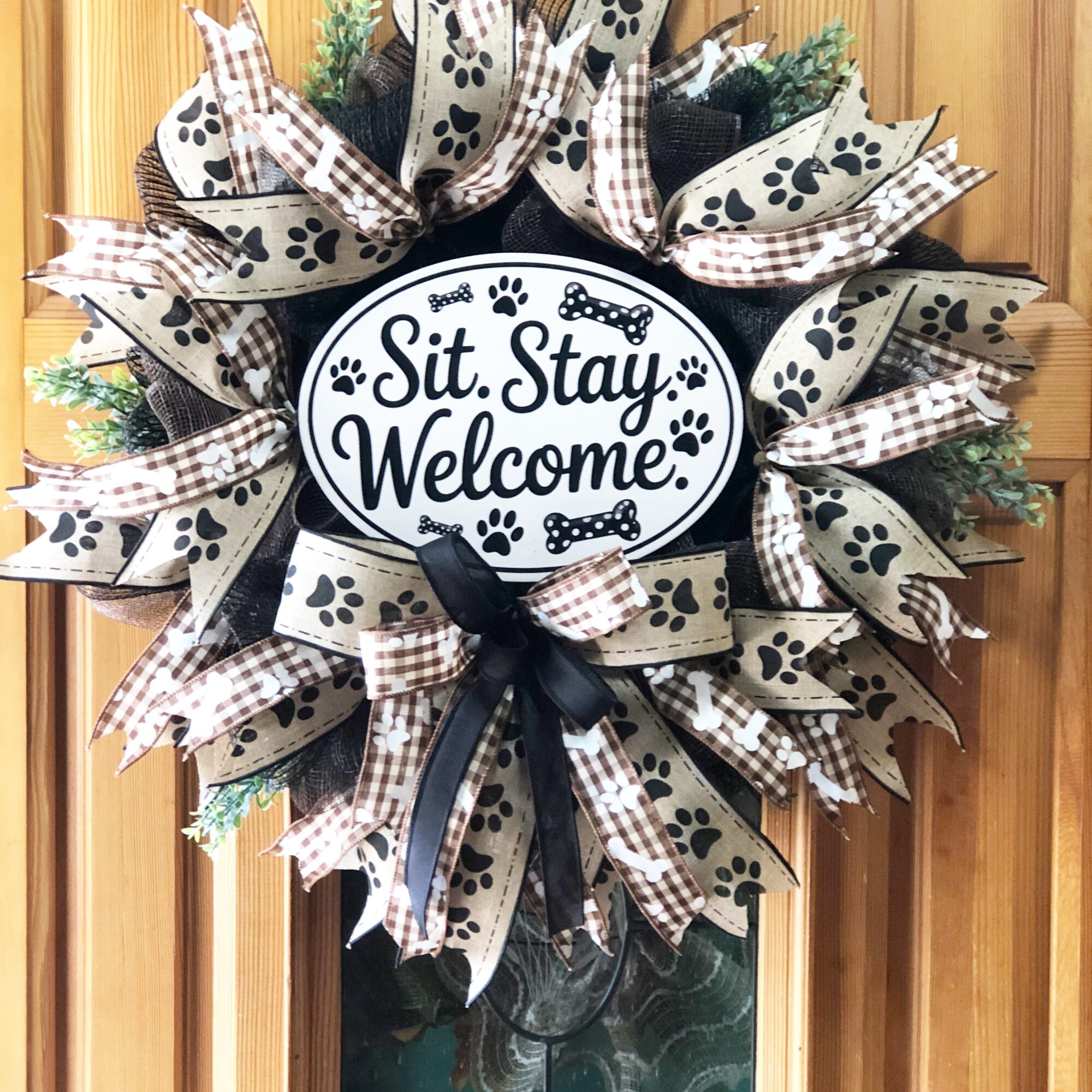 Decorative wreath with black, white, and checkered ribbons, featuring a central sign that says 'Sit. Stay. Welcome!'.