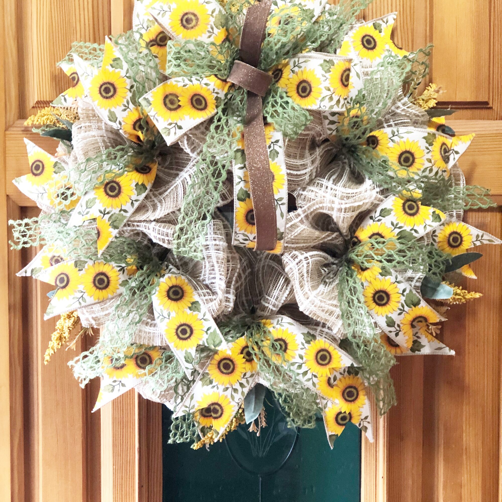 Decorative wreath with sunflower-themed ribbons, greenery, and a brown ribbon bow, hanging on a wooden door.