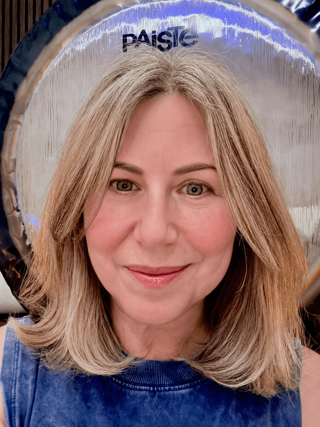 Woman with shoulder-length blonde hair smiling, standing in front of a reflective surface with the word 'PAISÉ' visible.