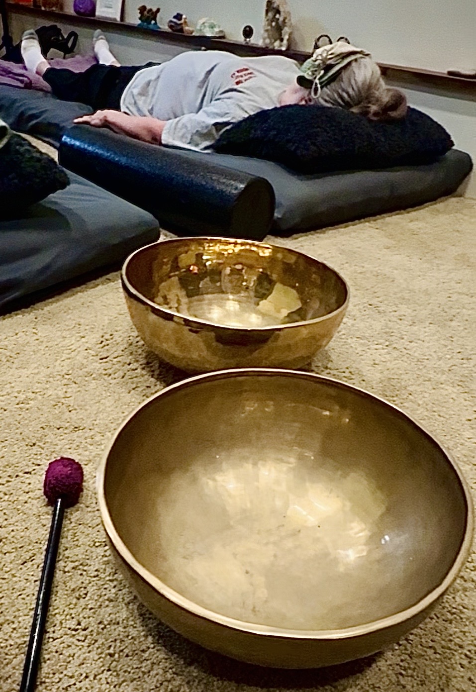 Person lying on a black mat with a pillow, in a room with a beige carpet, two large brass singing bowls in foreground.