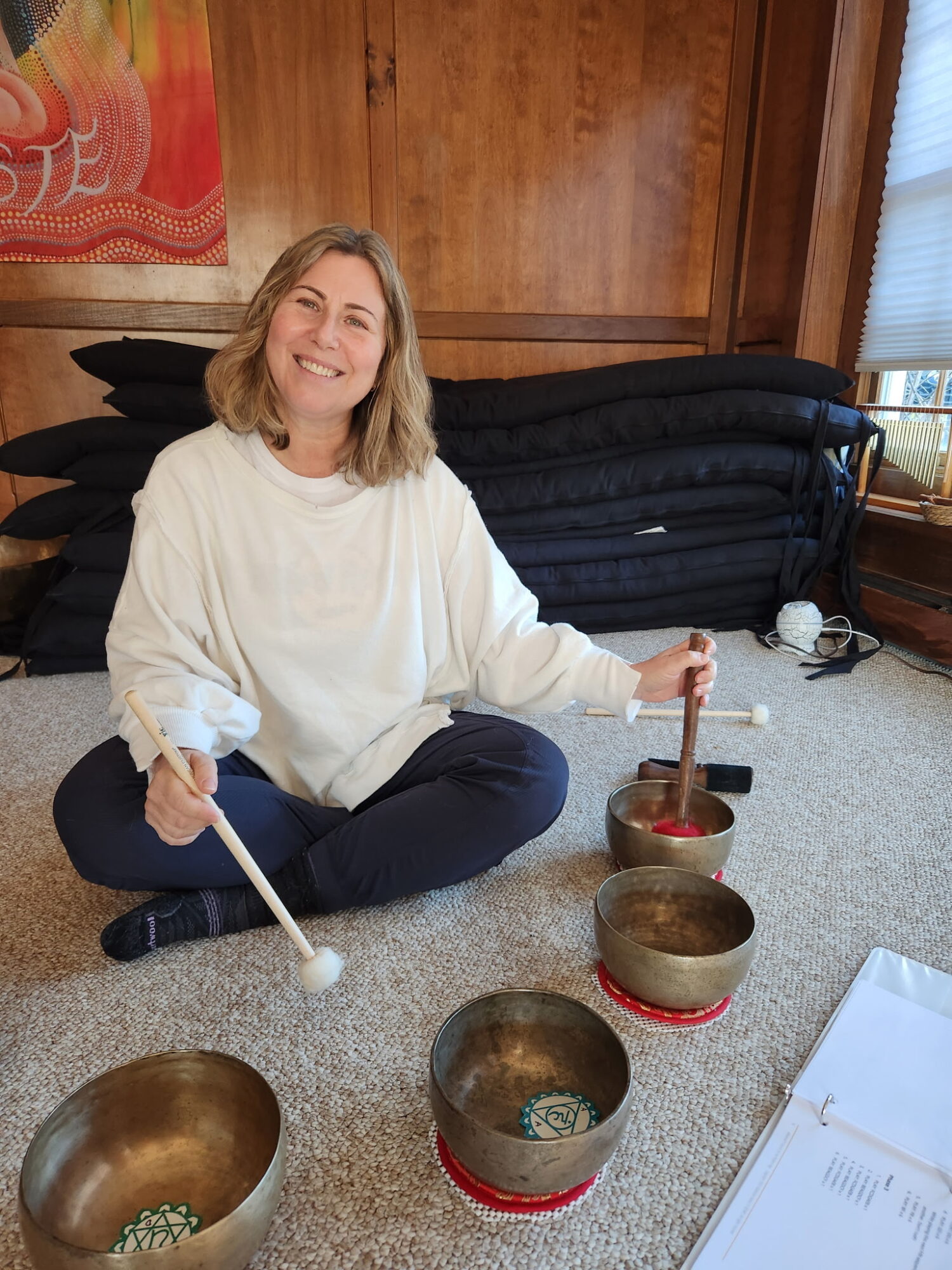 Woman sitting on carpeted floor, smiling, surrounded by singing bowls, in a room with wooden walls and window blinds.