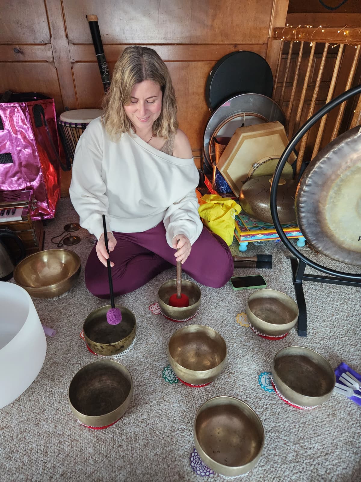 Young girl sitting on carpeted floor, surrounded by singing bowls, holding a mallet, in a room with wooden walls.