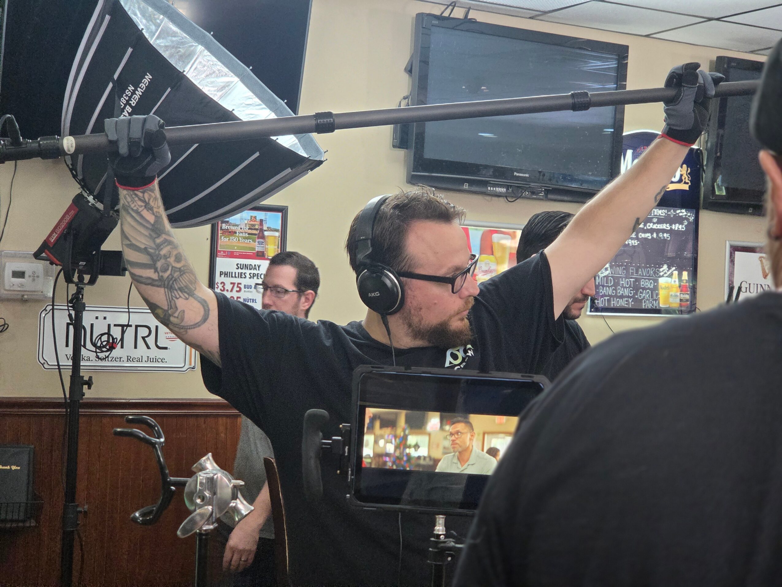 Man with headphones and tattoos holds a boom microphone overhead in a room with monitors and posters.