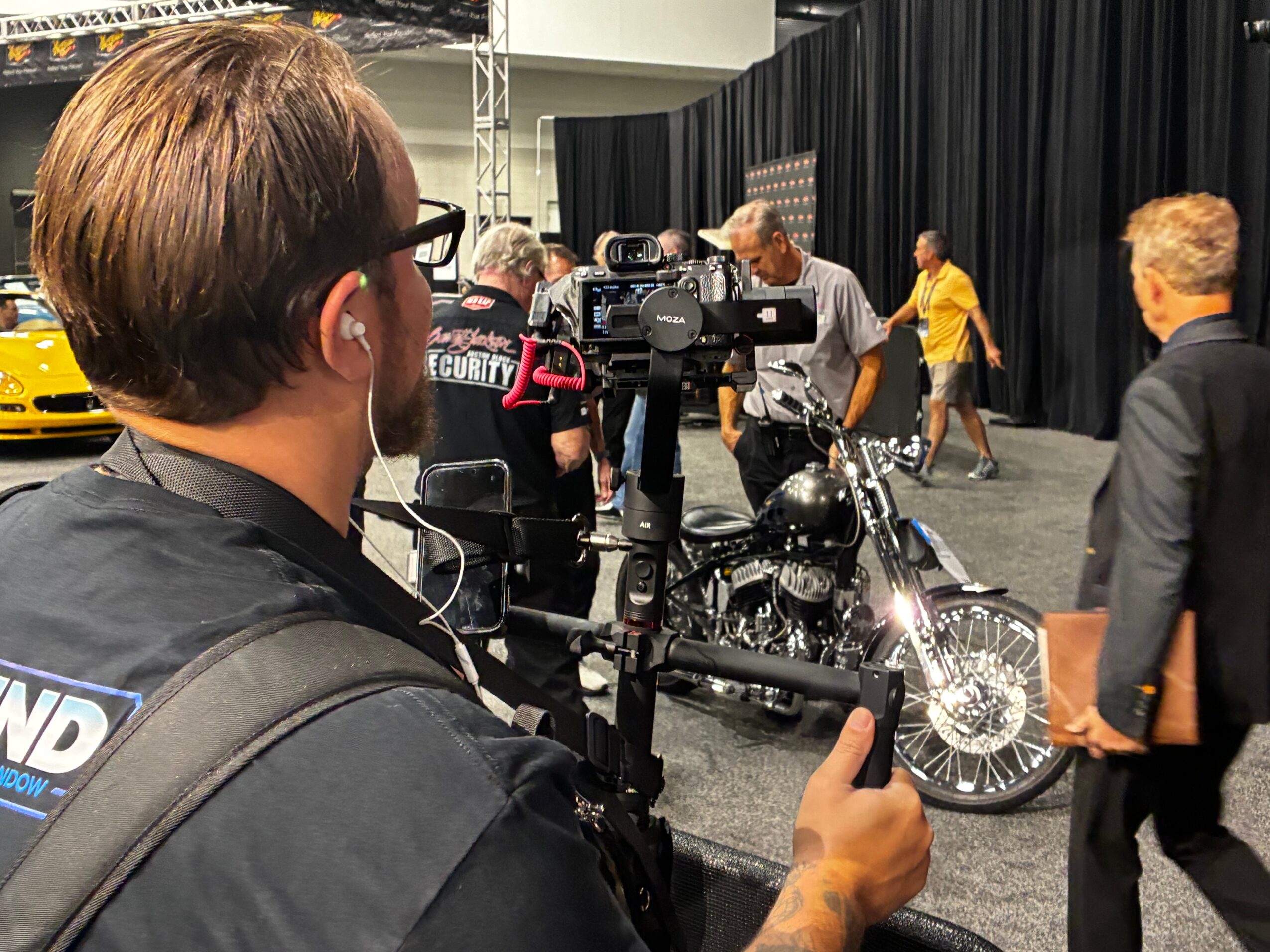 Person filming a motorcycle at an indoor event with people walking around and black curtains in the background.