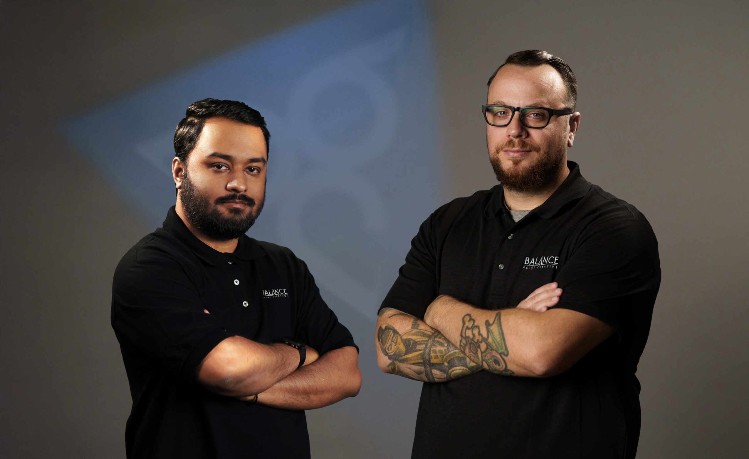 Two men with arms crossed, standing side by side in front of a gray background, wearing black shirts with logos.